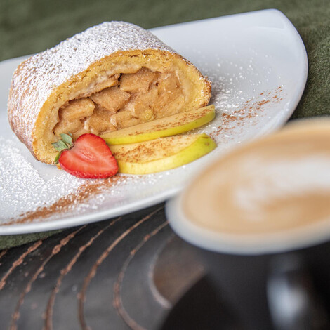 Apfelstrudel mit Puderzucker, Apfelscheiben und Erdbeere auf weißem Teller neben Tasse mit Kaffee.