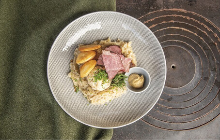 Teller mit einem Knödel, Surfleisch, Kraut und Scheiben von rosa gebratenem Fleisch. Separate kleine Schüssel mit einem Klecks Senf. Auf einem grauen Teller mit grünem Stoff und altem Herd als Hintergrund.