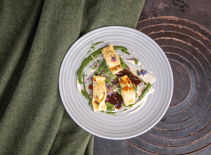 Drei mit Sauce und essbaren Blüten dekorierte Ravioli auf weißem Teller neben grünem Stoff und auf einer alten Herdplatte.