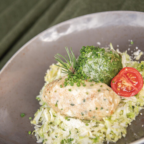 Ein Käseknödel und ein Spinatknödel auf einem Bett aus fein geschnittenem Kraut, garniert mit einem halbierten Kirschtomate, frischen Kräutern und geriebenem Käse auf einem grauen Teller.