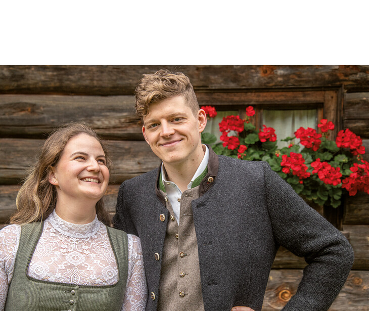 Zwei Personen in traditioneller Tracht vor einer Holzwand mit roten Blumen in einem Fenster.
