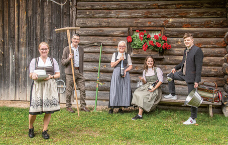 Five people in traditional dress in front of a wooden barn with flower boxes, one person holding a scythe, another holding a cooking pot with a wooden spoon, another holding a bottle, one sitting on a bench with glasses and a bottle in their hand, and one person standing in the foreground with plates and bowls.