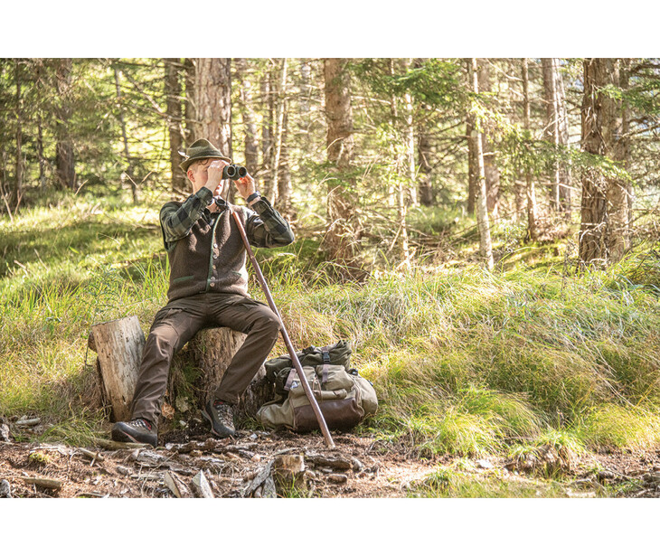 Person mit Jagdhut und Fernglas sitzt auf einem Baumstumpf im Wald neben Rucksack und Gewehr.