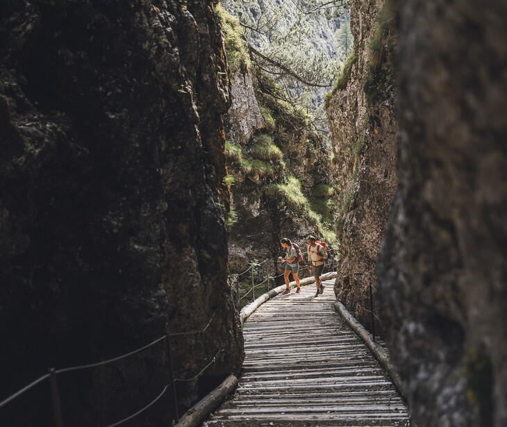 Two hikers with backpacks walking on a wooden path between tall rock walls in a canyon.