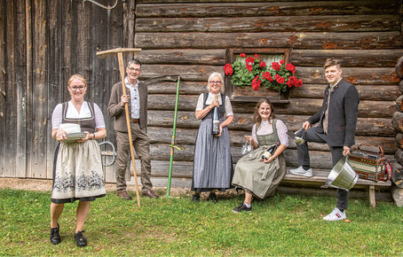 Fünf Personen in traditioneller Tracht vor einem Holzstadel mit Blumenkasten, eine Person hält eine Sense, eine andere einen Kochtopf mit Kochlöffel, eine weitere eine Flasche, eine sitzt auf einer Bank mit Gläser und Flasche in der Hand, eine Person steht im Vordergrund mit Tellern und Schüsseln.