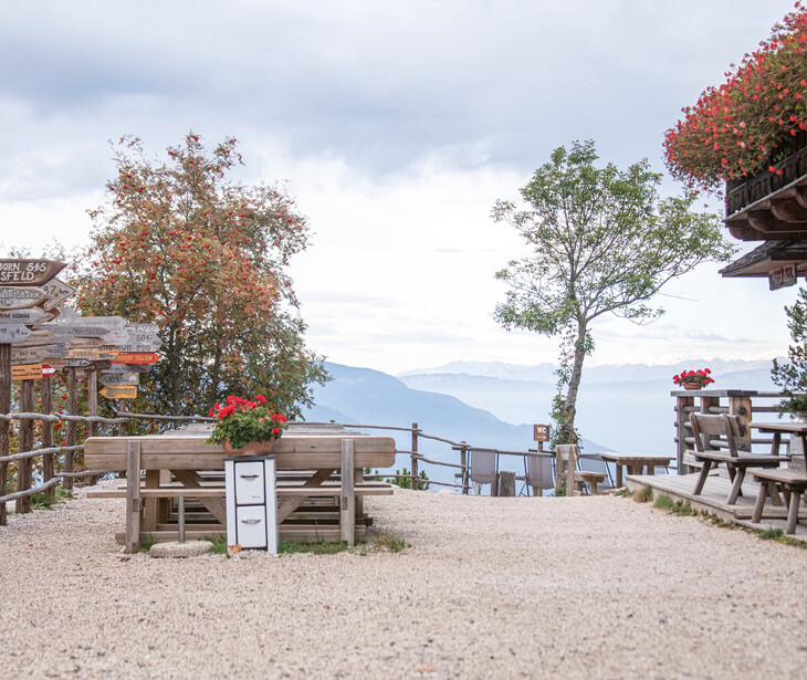 Terrasse mit Holzbänken und Tischen, Blumenkästen und Wegweisern vor Bergpanorama unter bewölktem Himmel.
