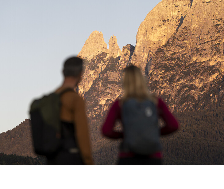 Zwei Personen mit Rucksäcken blicken auf eine Felsformation in den Bergen bei Sonnenlicht.
