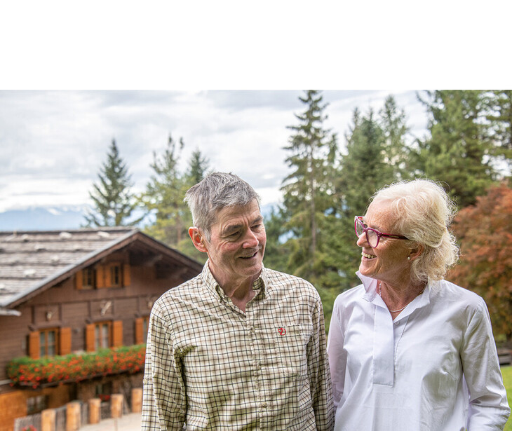 Älteres Paar steht vor einem Holzhaus mit Blumen in einem bewaldeten Berggebiet.