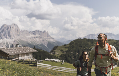 Two hikers with backpacks on a mountain trail with alpine huts and mountain scenery in the background.