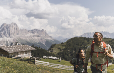 Zwei Wanderer mit Rucksäcken auf einem Bergweg mit Almgebäuden und Bergpanorama im Hintergrund.
