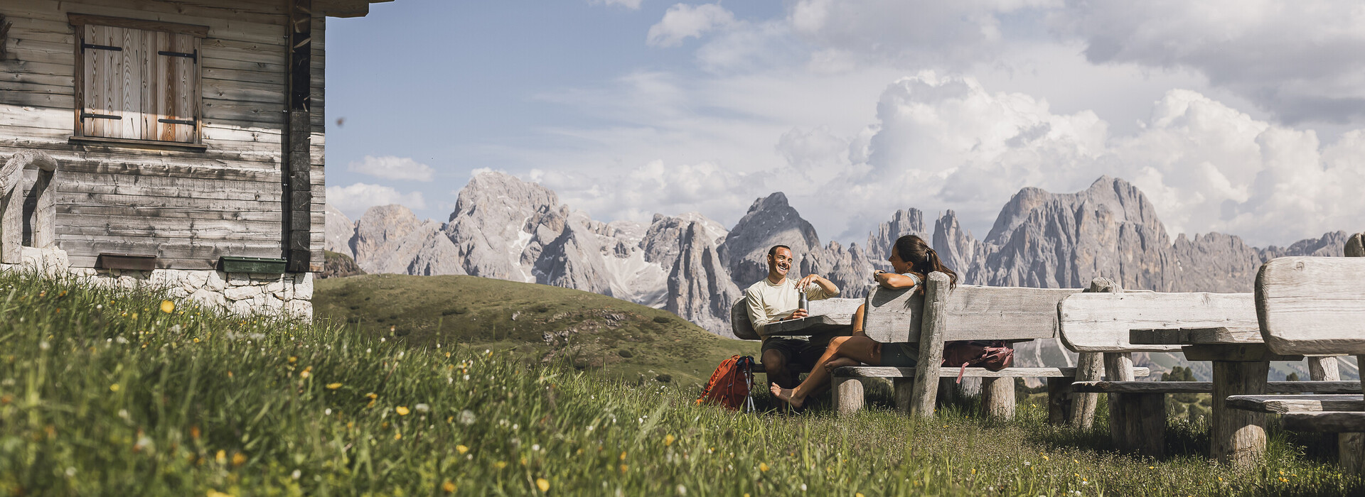 Two people sitting at a wooden table on a grassy meadow next to a wooden cabin with mountain peaks in the background.