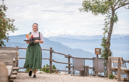 Kellnerin in traditioneller Tracht trägt ein Tablett mit Getränken auf einer Terrasse mit herrlichem Bergpanorama im Hintergrund.
