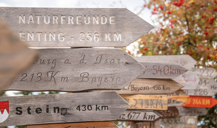 Mehrere hölzerne Wegweiser mit Entfernungsangaben zu verschiedenen Orten, unscharfer Hintergrund mit Bäumen.