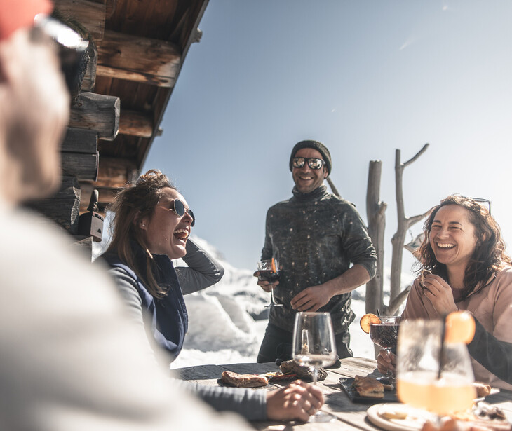 Un gruppo di quattro persone è seduto a un tavolo di legno all'aperto davanti a un rifugio di montagna con la neve sullo sfondo e beve da bicchieri.