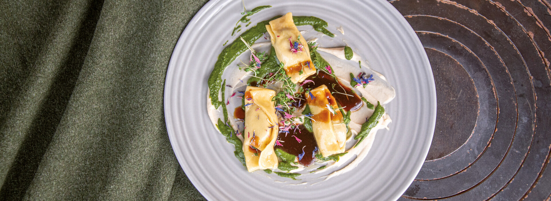 Drei mit Sauce und essbaren Blüten dekorierte Ravioli auf weißem Teller neben grünem Stoff und auf einer alten Herdplatte.
