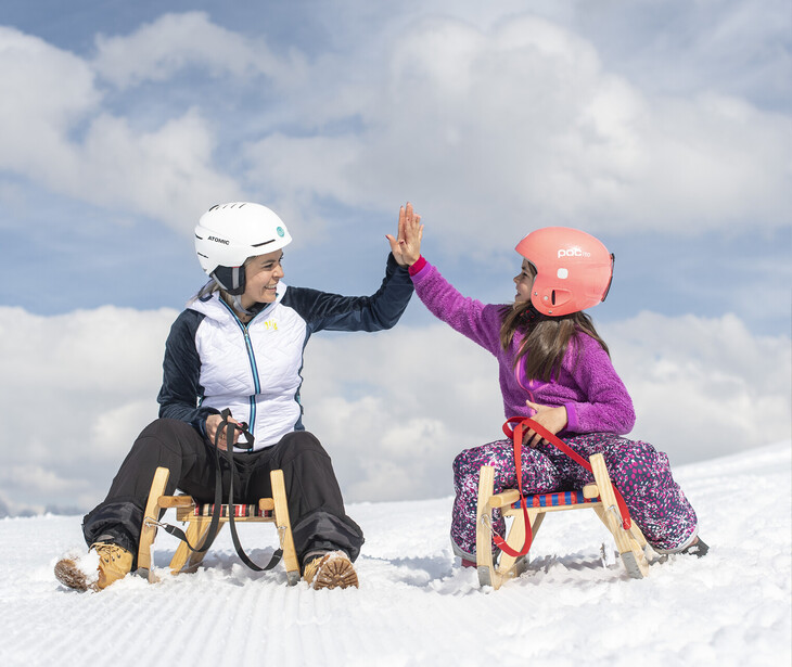 Eine Mutter und ihre Tochter sitzen mit Helm auf Holzschlitten im Schnee und geben sich einen High-Five.