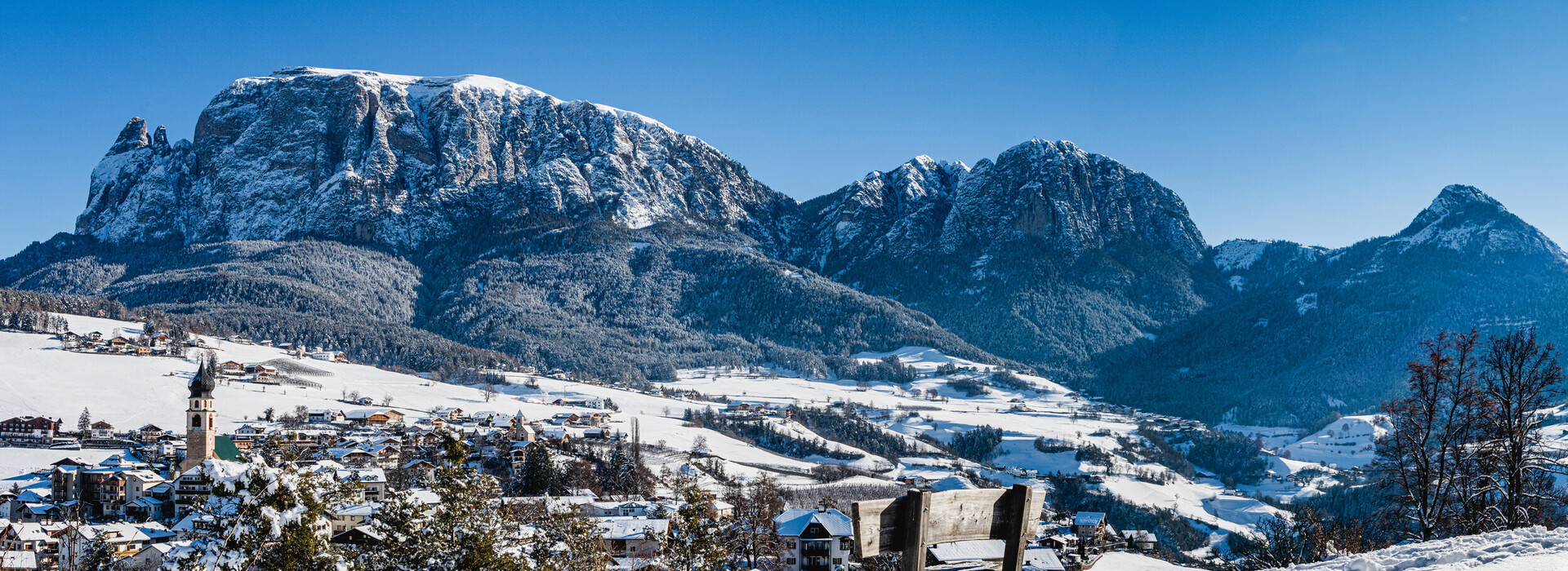 Panchina innevata su una collina con vista sul paesaggio montano innevato e sul villaggio sotto il cielo blu.