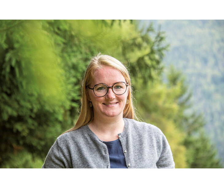 Frau mit langen blonden Haaren trägt eine graue Strickjacke über einem dunkelblauen Oberteil vor bewaldetem Hintergrund.