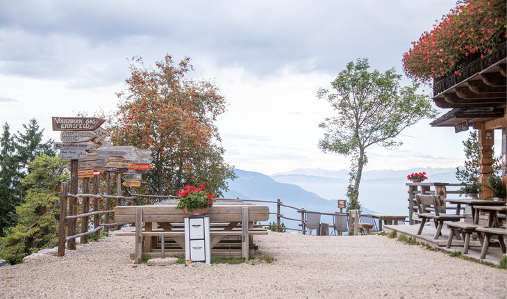 Terrace with wooden tables and benches, flower boxes, and signposts against a mountain panorama and cloudy sky.