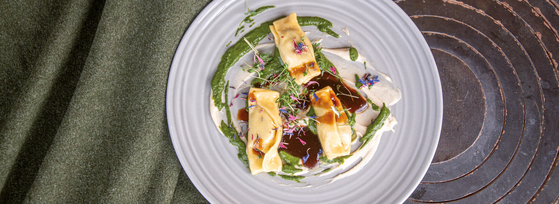 Three ravioli decorated with sauce and edible flowers on a white plate next to green fabric and on an old stove top.