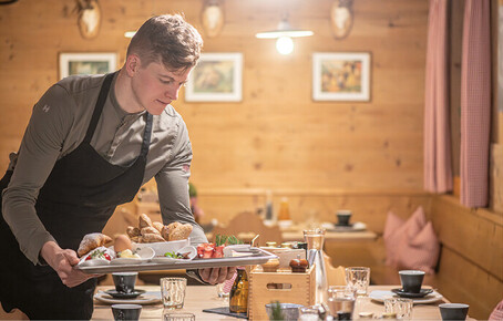 Kellner mit schwarzer Schürze trägt Tablett mit Frühstück in rustikalem Gastraum mit Holzwänden und gedecktem Tisch.