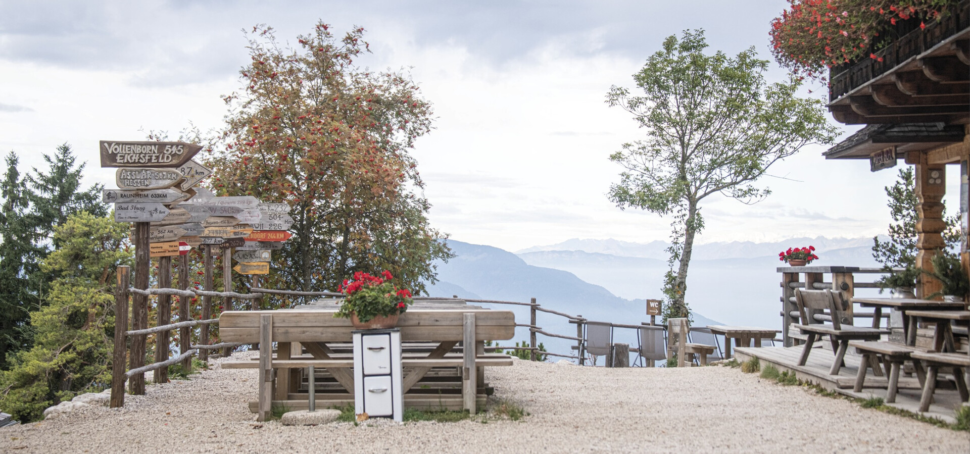 Terrasse mit Holztischen und Bänken, Blumenkästen und Wegweisern vor Bergpanorama und bewölktem Himmel