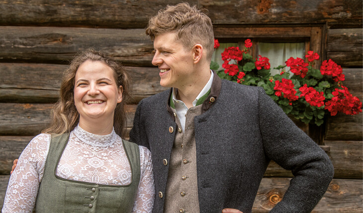 Two people in traditional attire standing in front of a wooden wall with red flowers in the window.