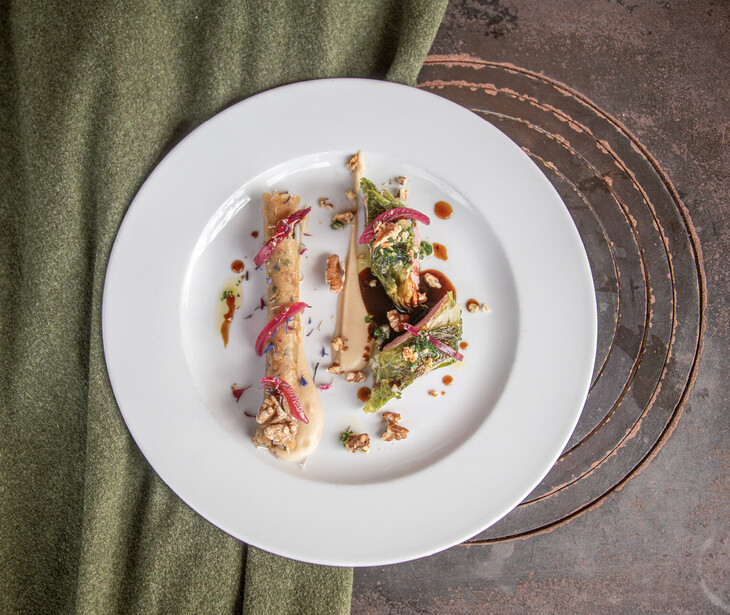 Weißer Teller mit kunstvoll angerichteter Speise, garniert mit roten Zwiebeln und Nüssen, auf grauem Untergrund neben grünem Stoff.