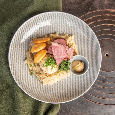 Piatto con un canederlo, carne affumicata, crauti e fette di carne rosata arrosto. Piccola ciotola separata con una cucchiaiata di senape. Su un piatto grigio con tessuto verde e vecchia stufa come sfondo.