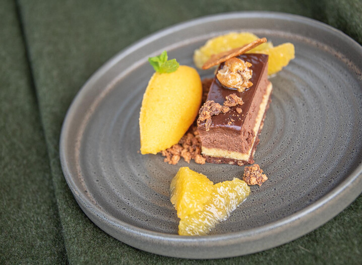 Schokoladendessert mit glänzender Glasur, garniert mit Nuss und Krokant, dazu eine Kugel Sorbet und Fruchtstücke auf grauem Teller.