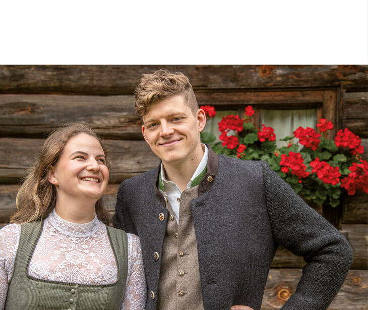 Zwei Personen in traditioneller Tracht vor einer Holzwand mit roten Blumen in einem Fenster.