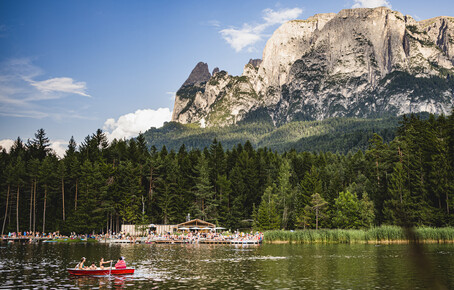 Bergsee mit Tretbootfahrern, bewaldetem Ufer und markantem Felsmassiv im Hintergrund.