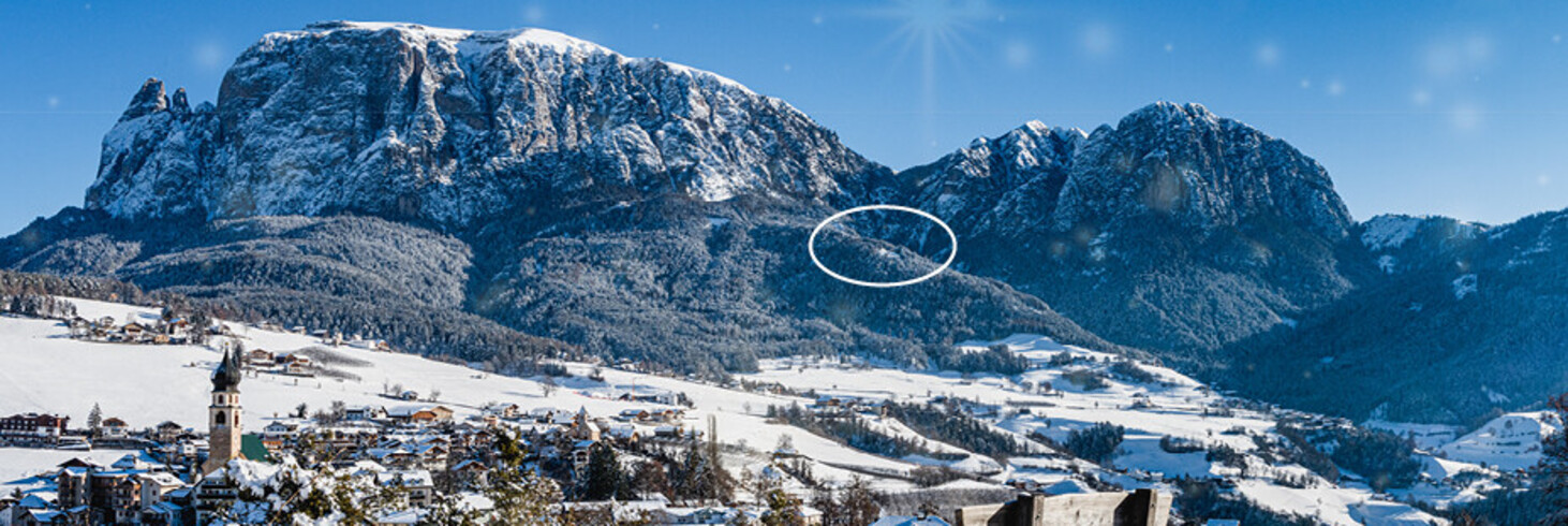 Schneebedeckte Berglandschaft mit Dorf im Tal und Holzbänken im Vordergrund unter klarem blauem Himmel.