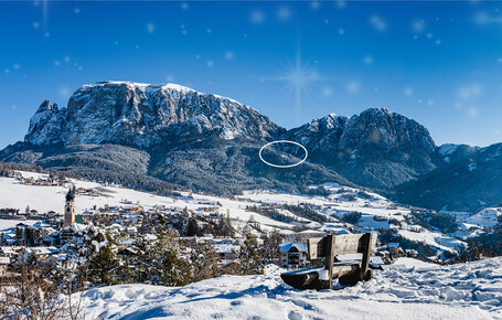 Schneebedeckte Berglandschaft mit Dorf im Tal und Holzbänken im Vordergrund unter klarem blauem Himmel.