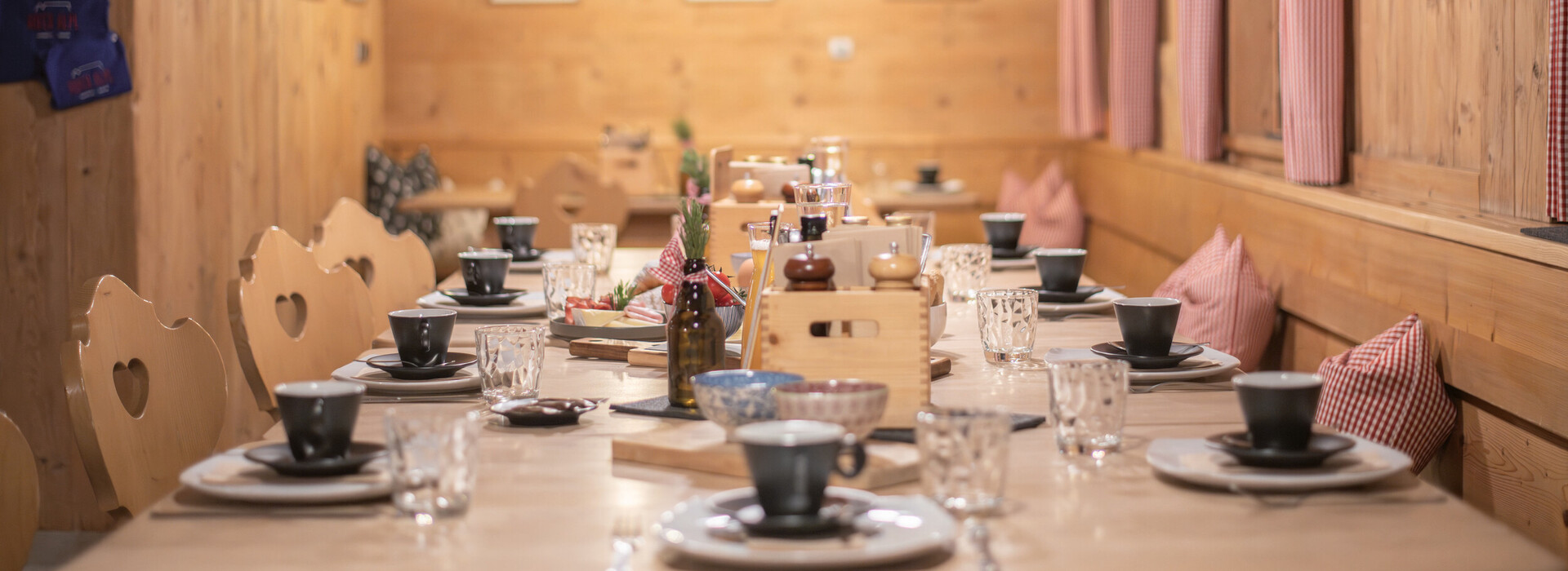 Wood-paneled room with long set dining table, wooden chairs with heart-shaped cutouts, and red-and-white checkered curtains.