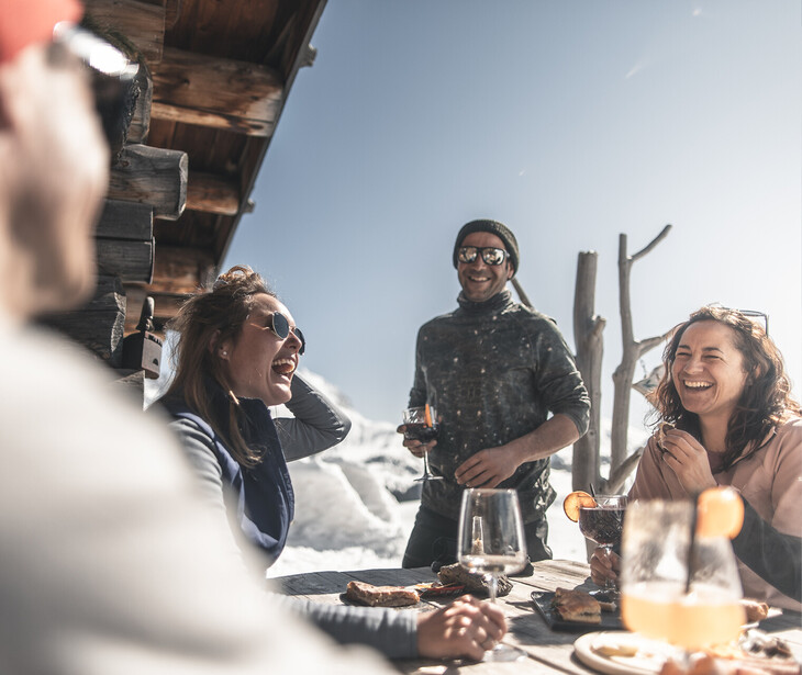 Gruppe von vier Personen sitzt an einem Holztisch im Freien vor einer Berghütte mit Schnee im Hintergrund und trinkt aus Gläsern.